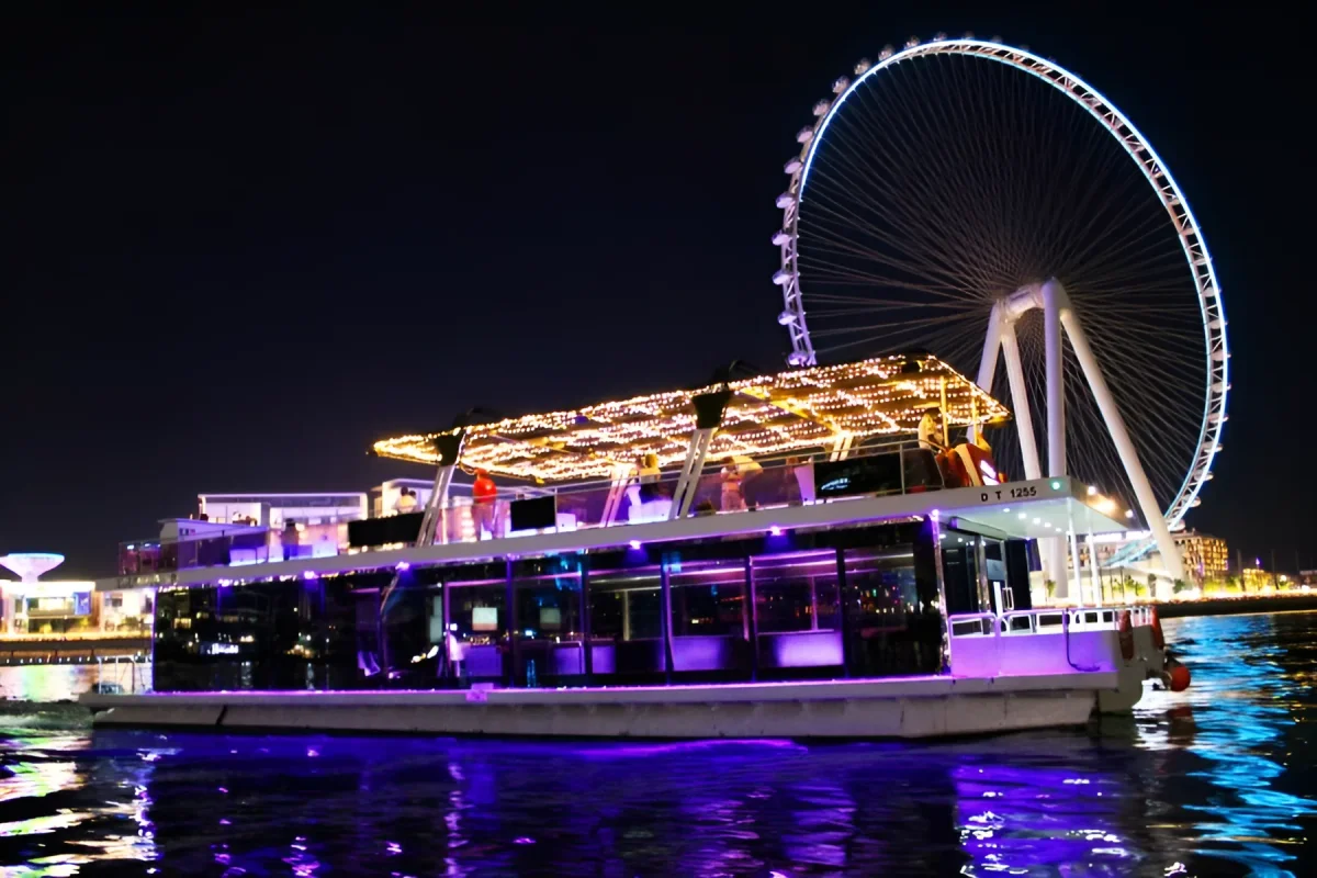Double-deck boat lit up with purple lighting cruising Dubai Marina at night, Ain Dubai wheel glowing behind