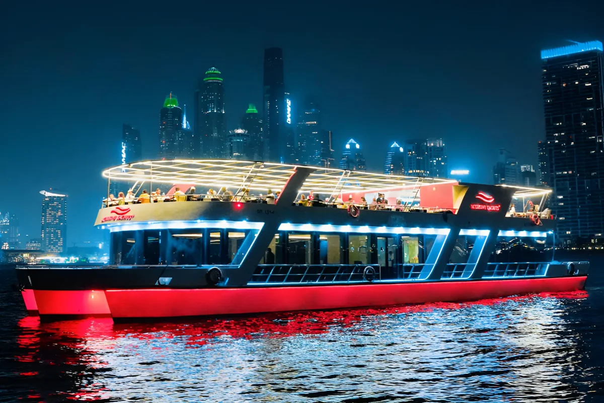 Houseboat lit up red and white cruising Dubai Marina at night with the skyline behind